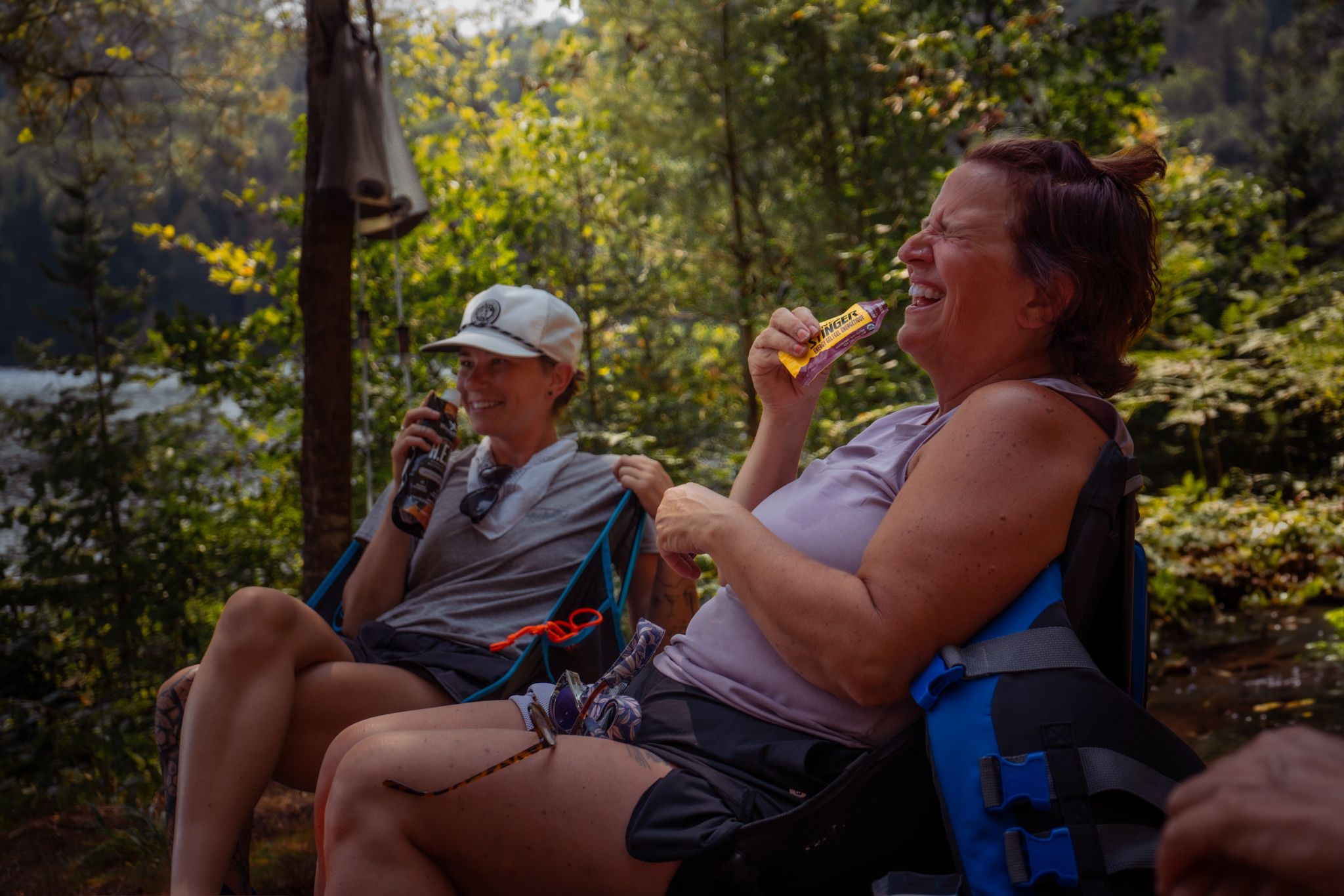 Two people sit in camping chairs enjoying beverages in a sunny forest clearing on a summer afternoon.