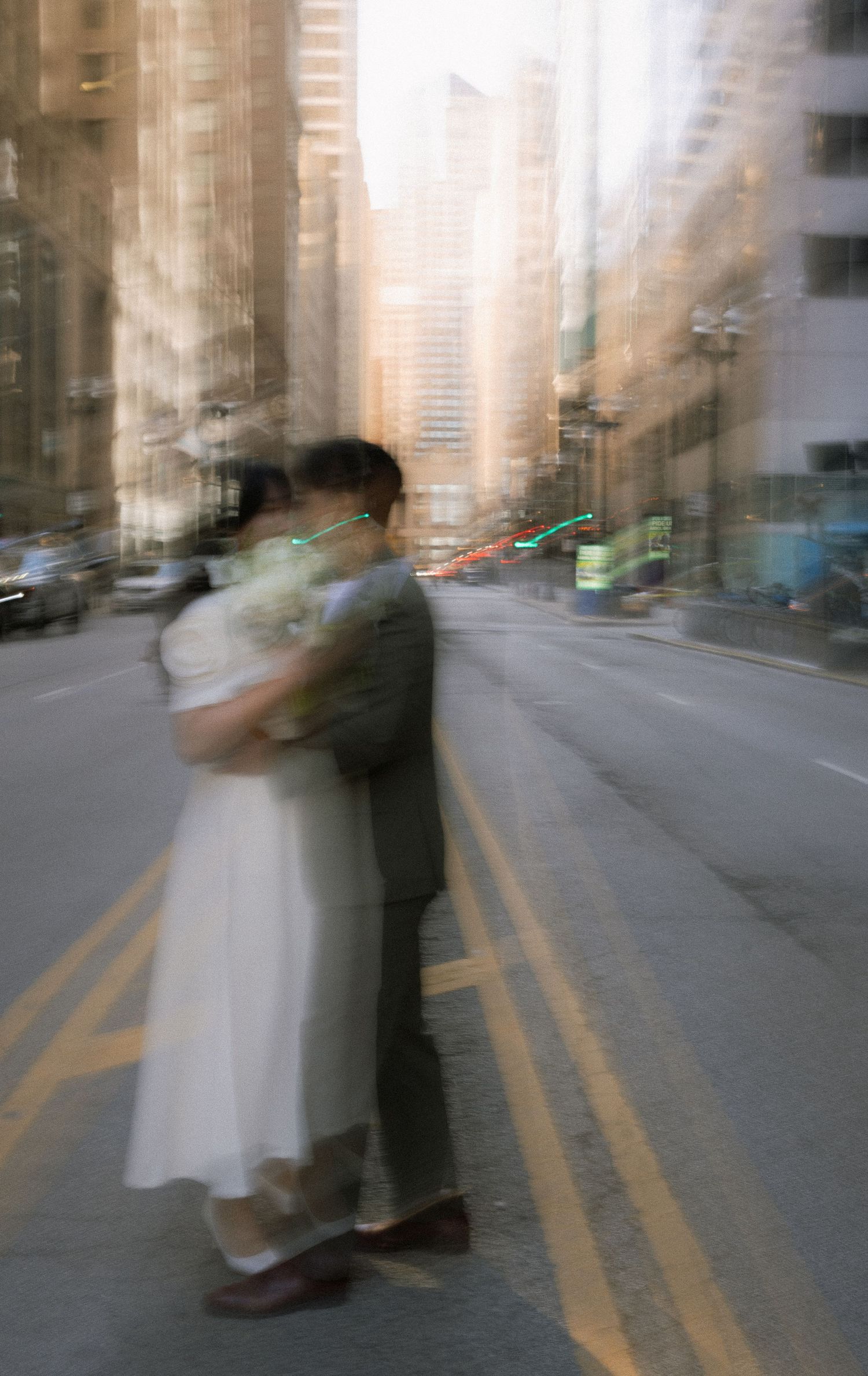 A blurred motion shot of two figures embracing in an urban street with tall buildings in the background.