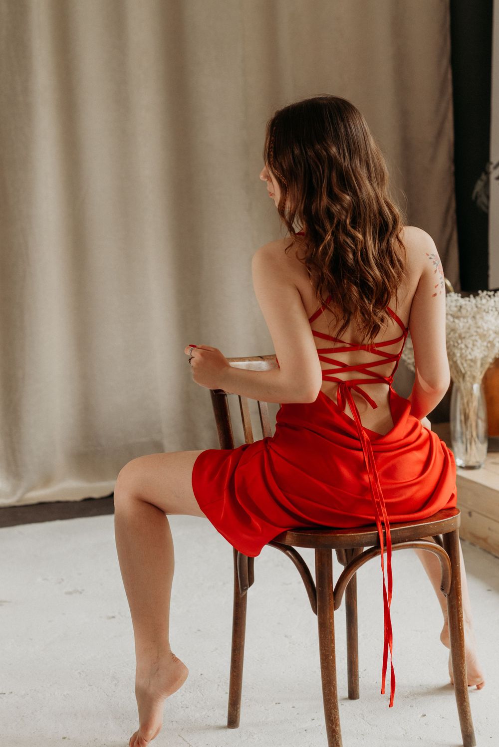 Figure in flowing red dress seated on wooden chair in window light.