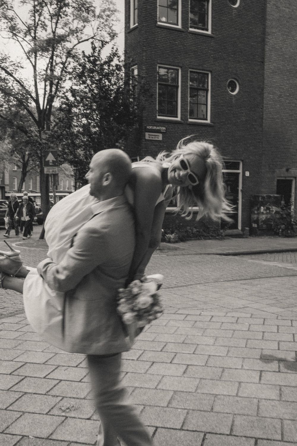 A bald man carries a laughing blonde woman on his back while walking on a cobblestone street in Amsterdam.