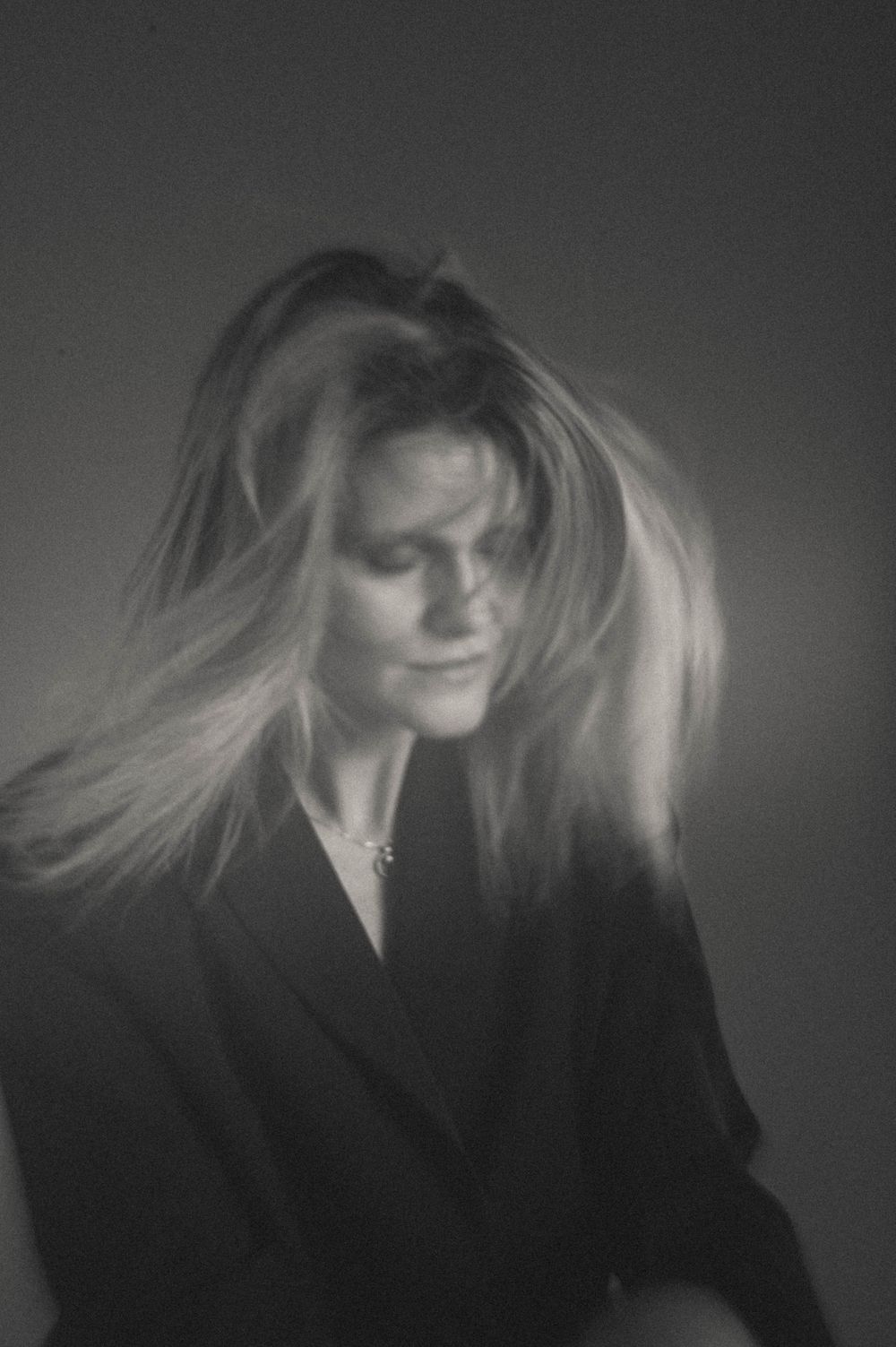 Black and white portrait of a woman with flowing blonde hair wearing a dark blazer, looking down dramatically.