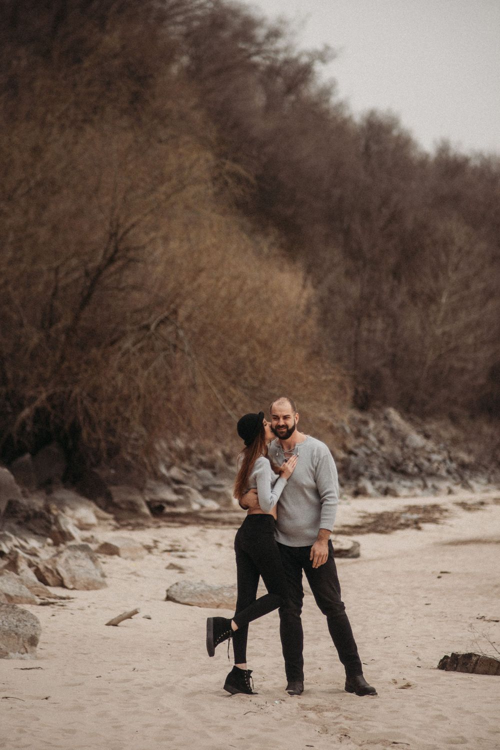 Playful couple interacting on sandy beach with winter brush in background.