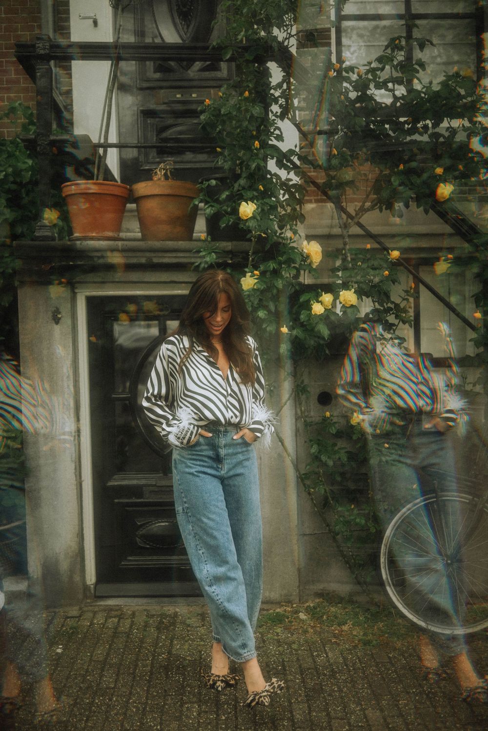 Woman in striped shirt and blue jeans poses outside a building with yellow roses and terracotta pots.