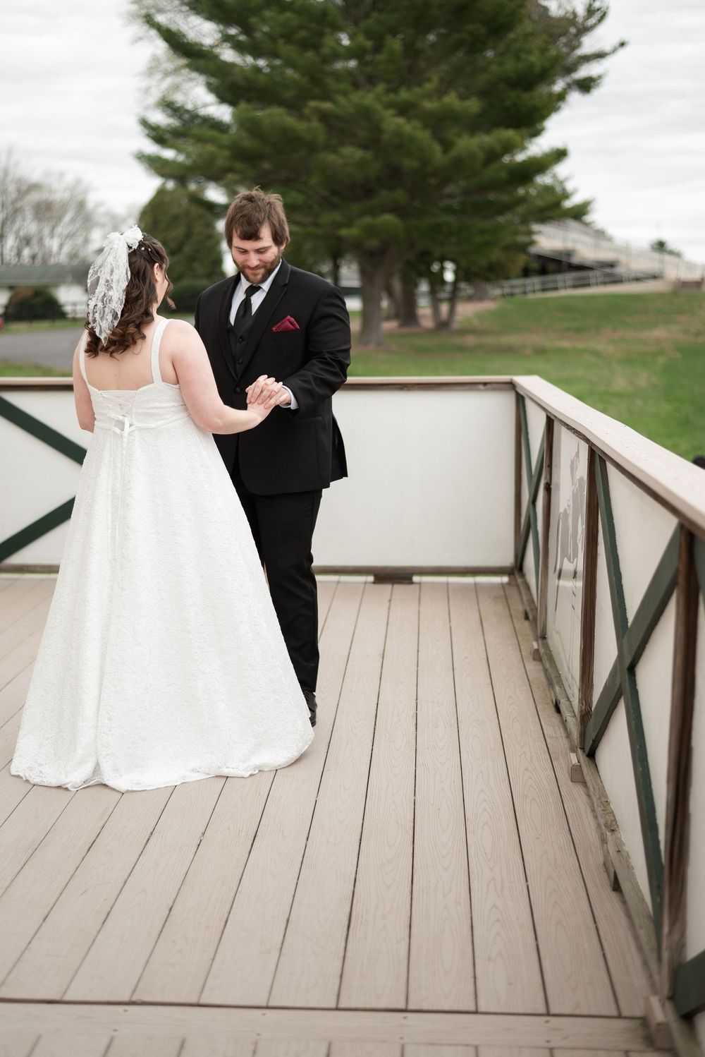 A couple in wedding attire embrace on a wooden deck with evergreen trees in the background on their special day.