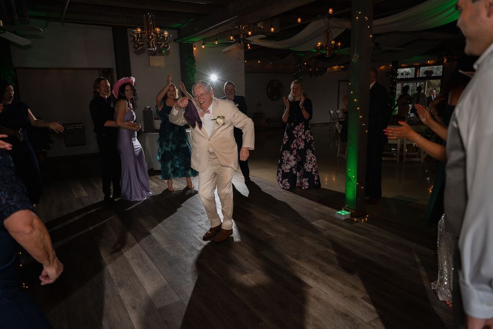 Groom in white suit dancing energetically on wedding reception dance floor surrounded by guests.