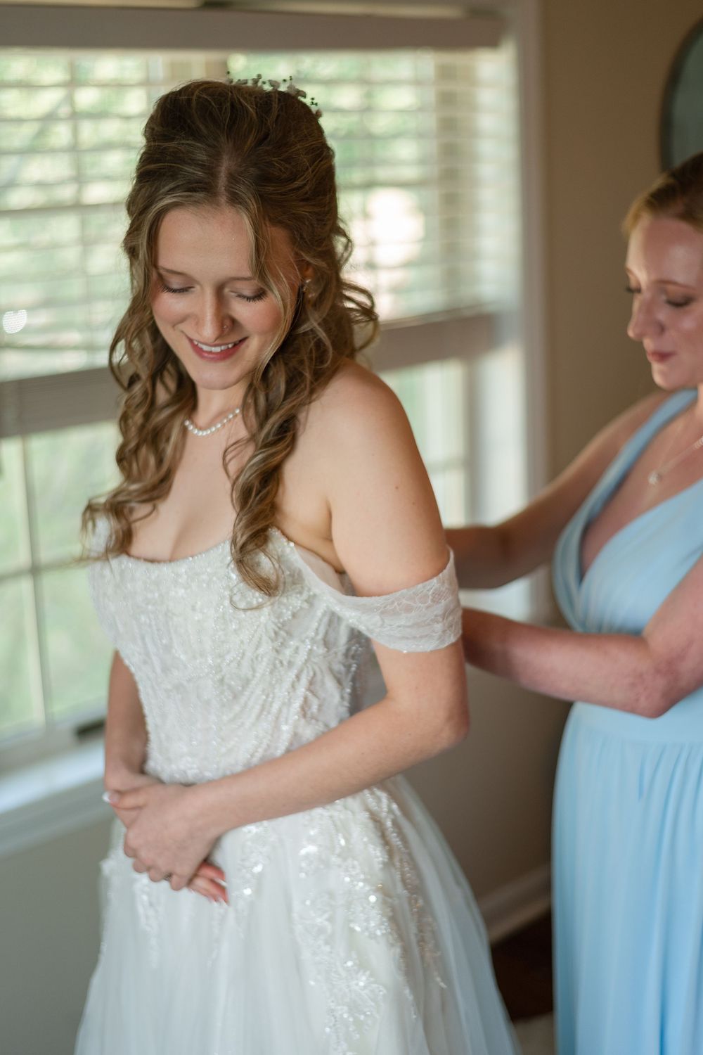 Bride in white off-shoulder gown smiles as bridesmaid in blue dress helps fasten her wedding dress near a window.