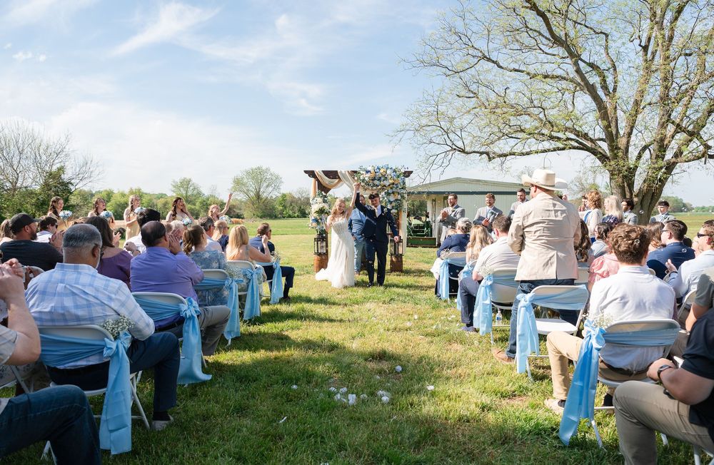 Outdoor wedding ceremony on green lawn with guests seated in chairs adorned with blue ribbons under a sunny sky.