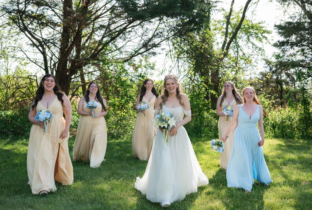 Bride in white gown with bridesmaids in champagne and blue dresses holding bouquets outdoors on green lawn.