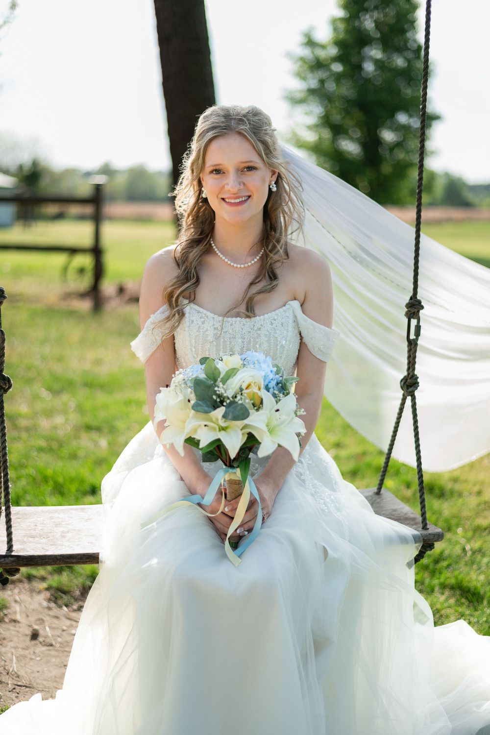Smiling bride in white off-shoulder gown sits on swing holding bouquet of lilies and hydrangeas with veil flowing.