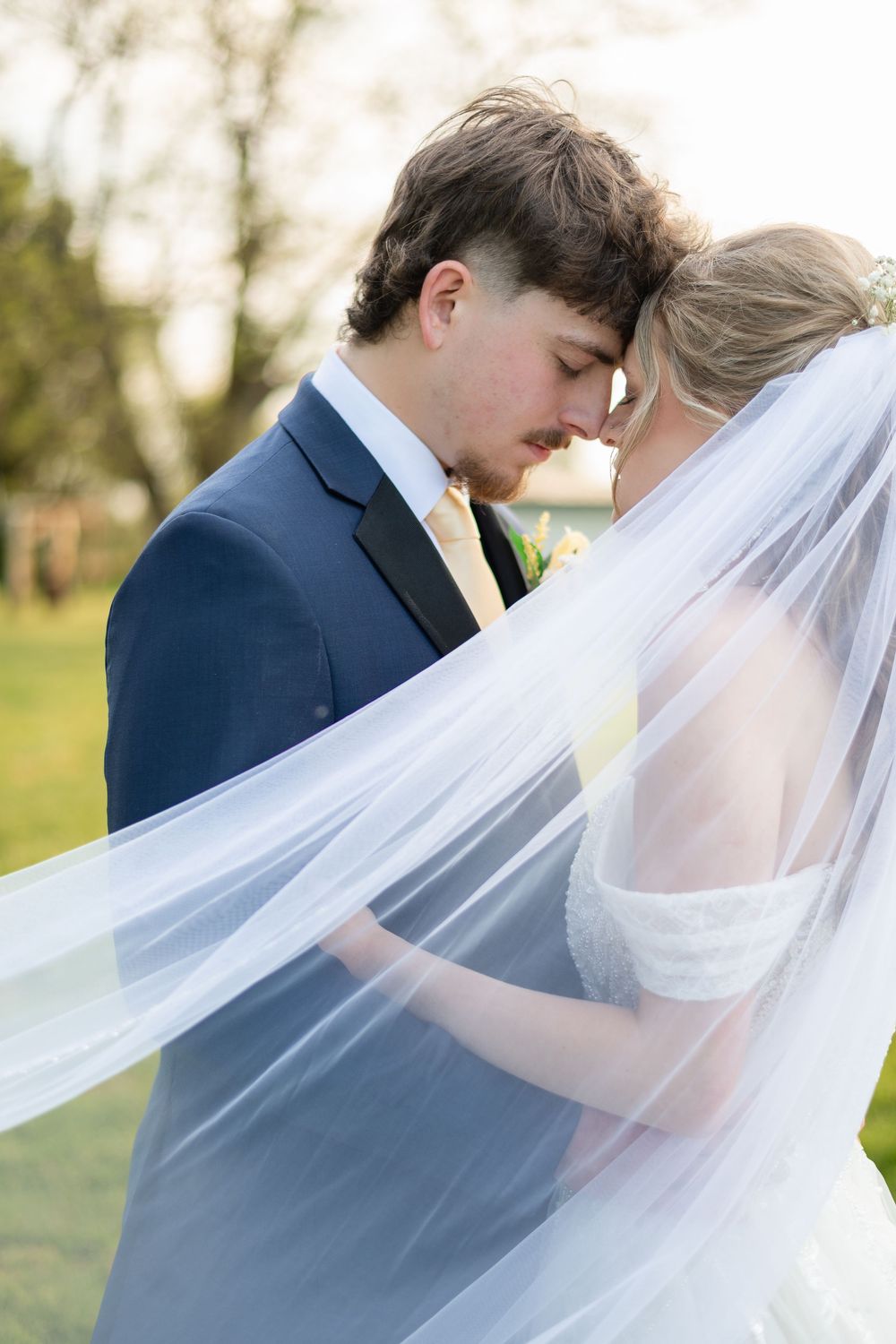 Bride and groom share intimate forehead touch under flowing veil outdoors in golden light.