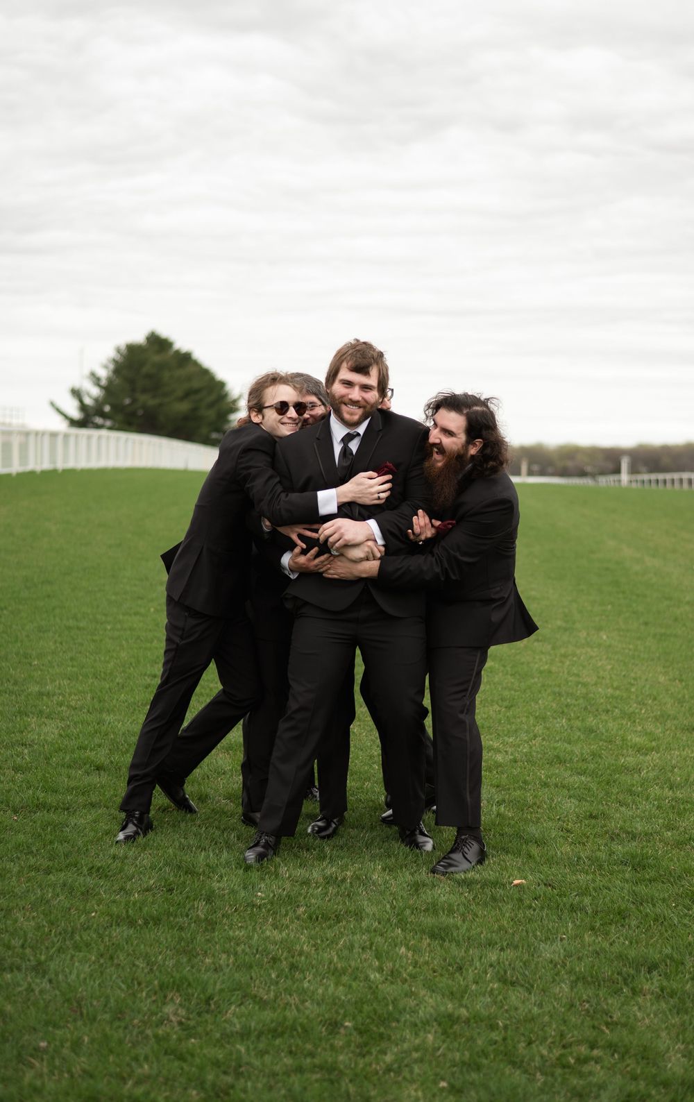 Three people in black suits playfully pose together on a green lawn with white fencing in the background.