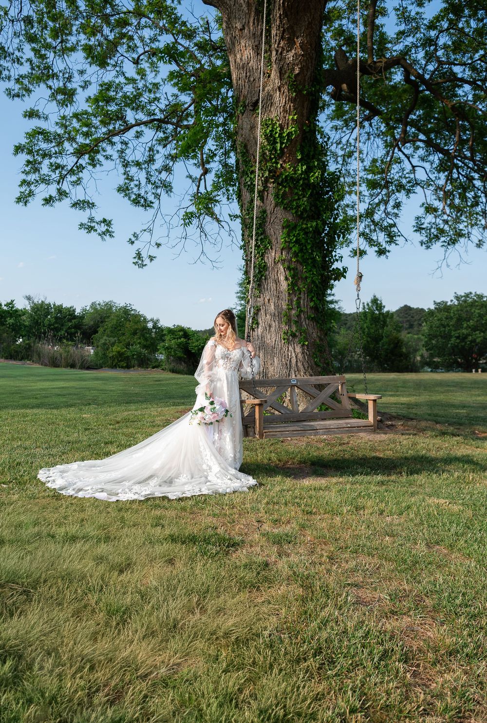 Bride in white lace gown with long train stands beside rustic bench under large oak tree on green lawn.