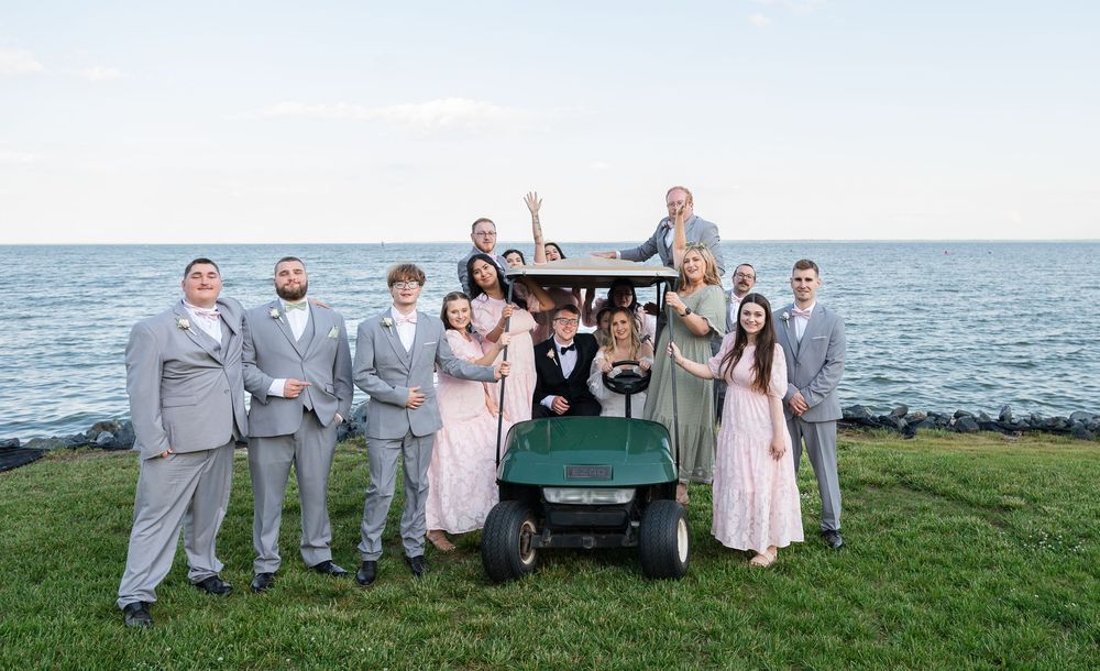 Wedding party poses with golf cart by waterfront, groomsmen in gray suits and bridesmaids in pink dresses.