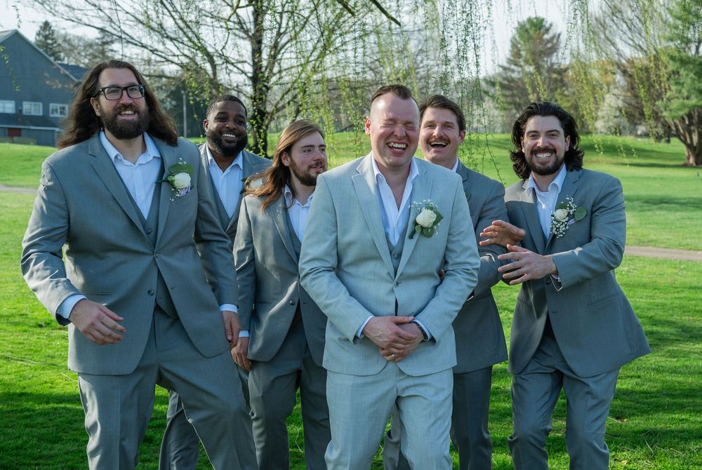 Groom and groomsmen laughing together outdoors in matching gray suits with boutonnieres on a sunny wedding day.