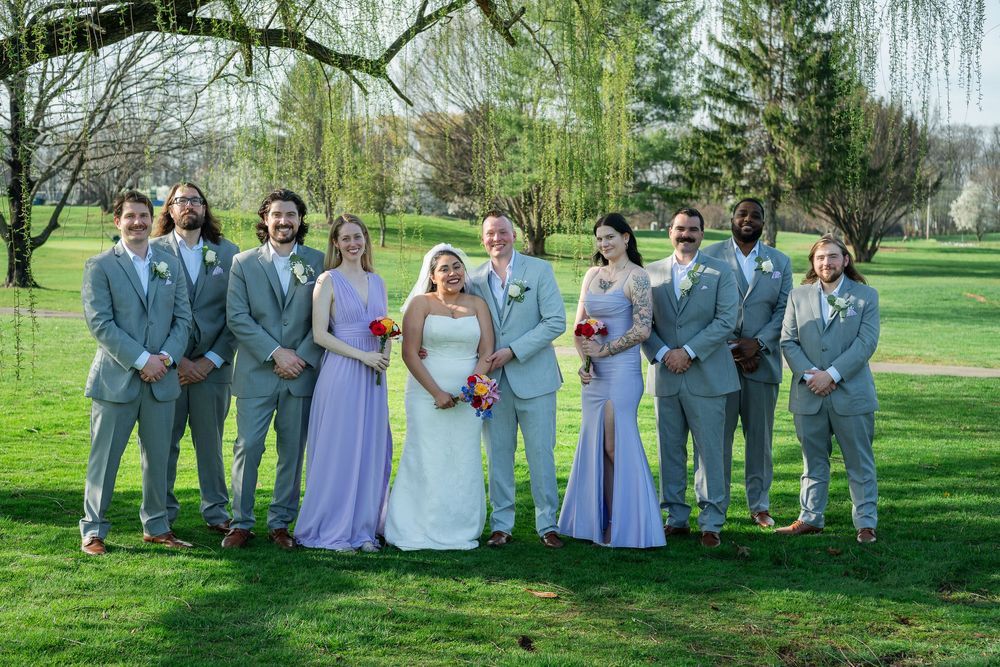 Wedding party poses outdoors in gray suits and lavender bridesmaid dresses with bride and groom centered on green lawn.