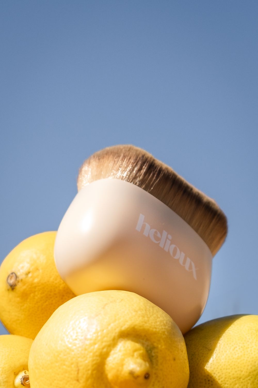Makeup brush with Helioux  branding rests on fresh yellow lemons against blue background.