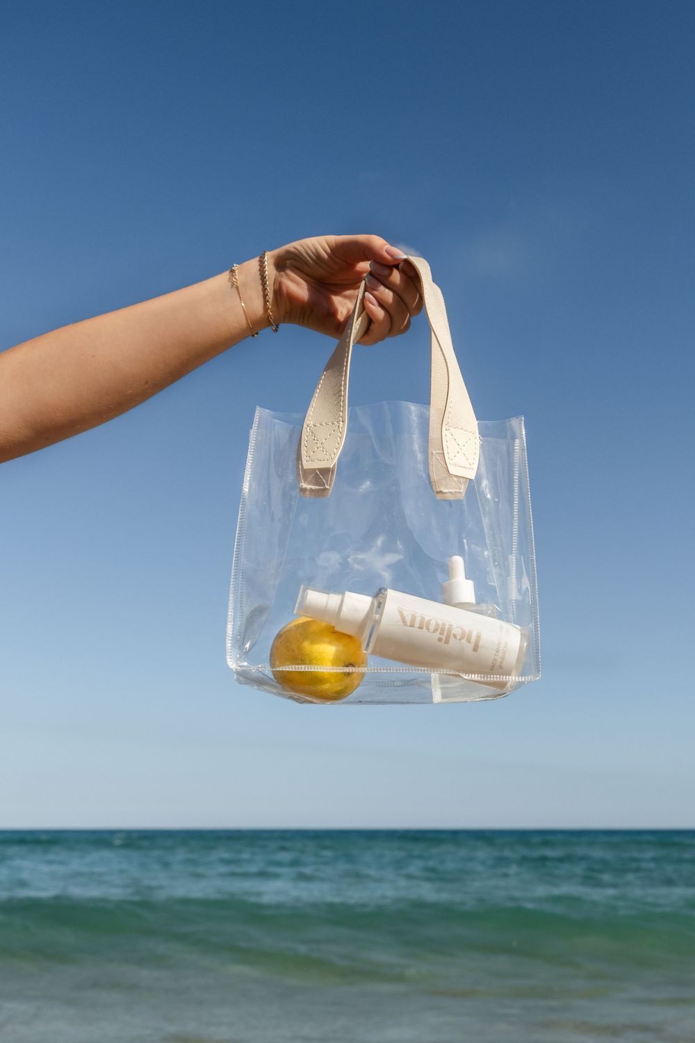 Hand holding clear beach tote bag with skincare products and citrus fruit against ocean backdrop.