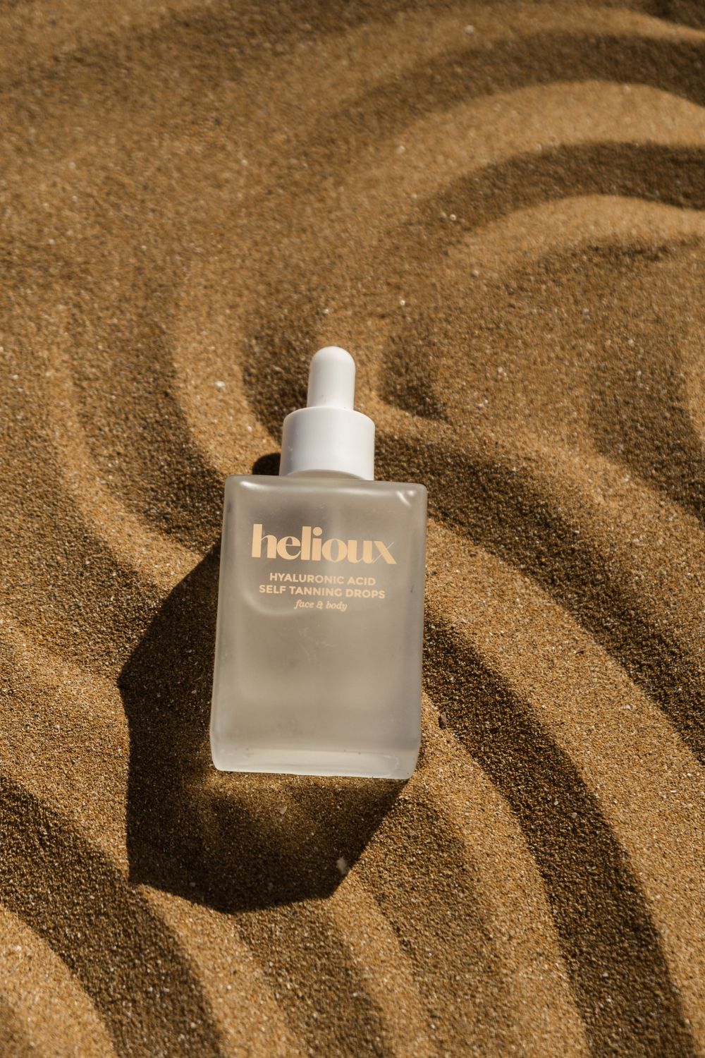 Glass skincare bottle with Helioux  branding placed artistically in textured sand.