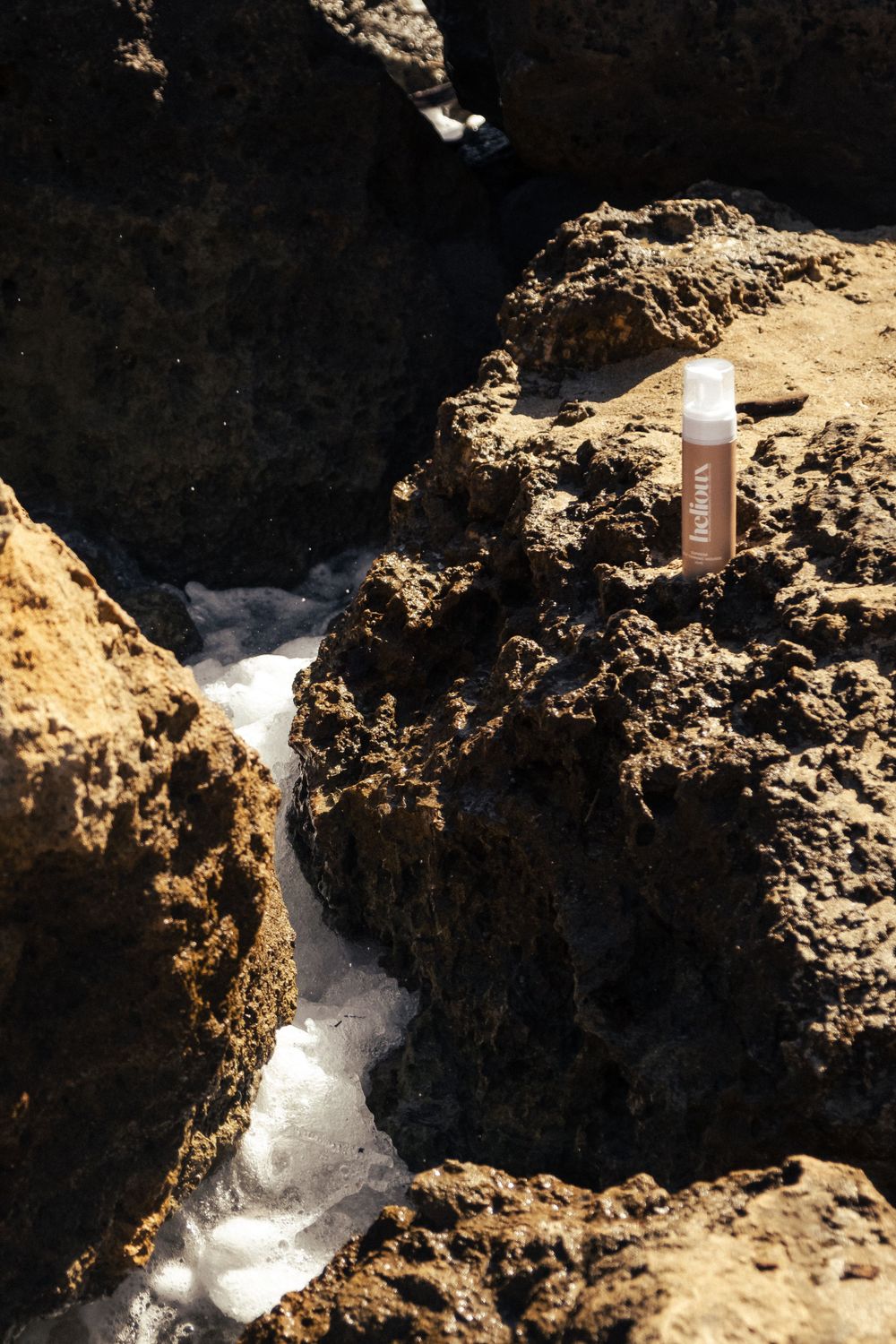 Skincare product bottle positioned near flowing water on rocky coastal formation.