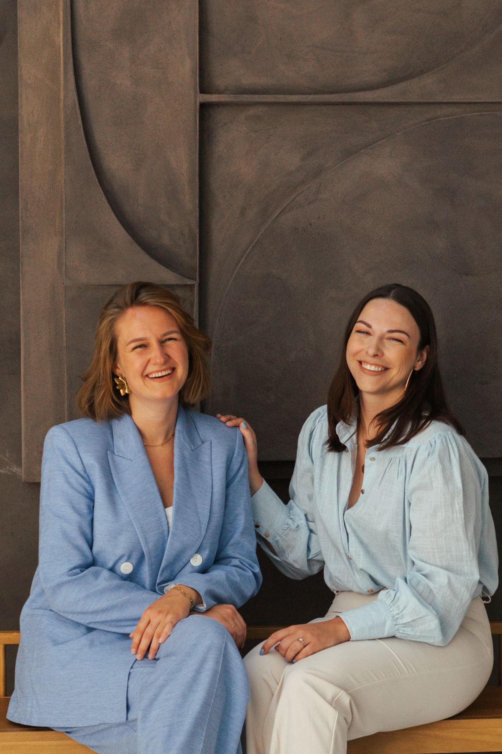 Two women in blue and white business attire share a candid moment while seated against a dark backdrop.