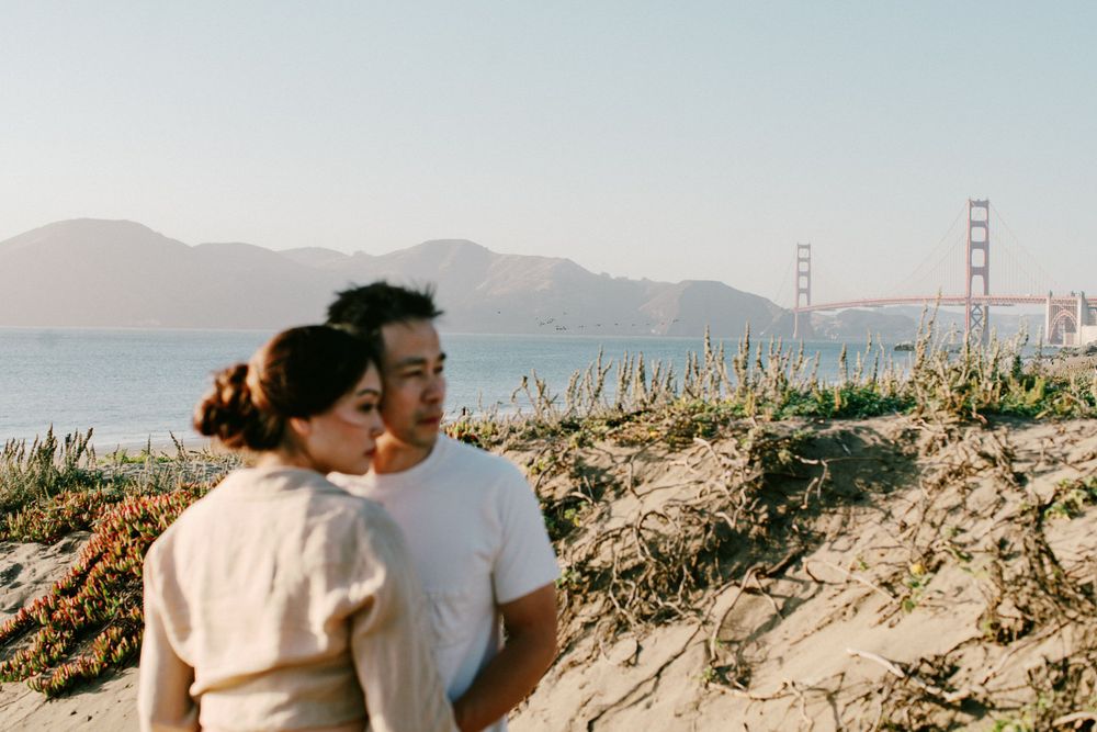 Romantic couple embracing at Baker Beach with the Golden Gate Bridge visible in the background.