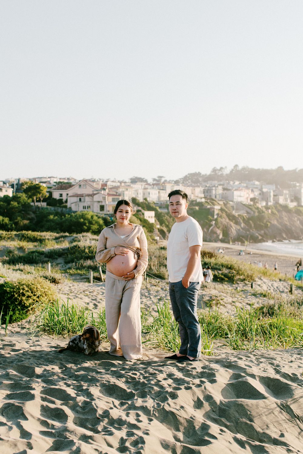 A couple and their dog posing together on a sandy beach with coastal homes in the background.