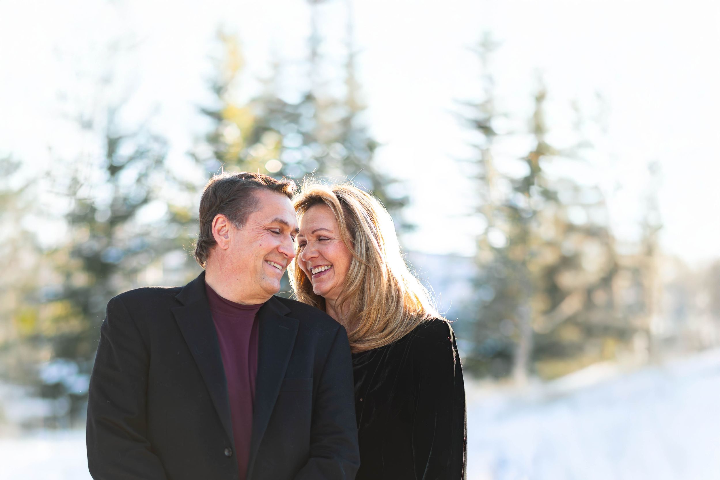 Winter portrait of couple in warm clothing against snowy evergreen backdrop.