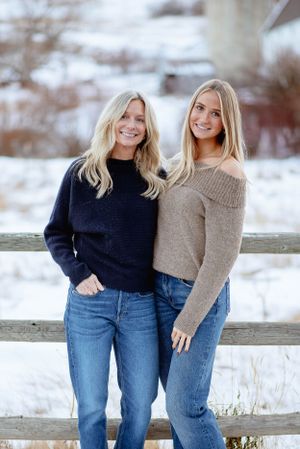 Two people in winter sweaters leaning against wooden fence in snowy setting.