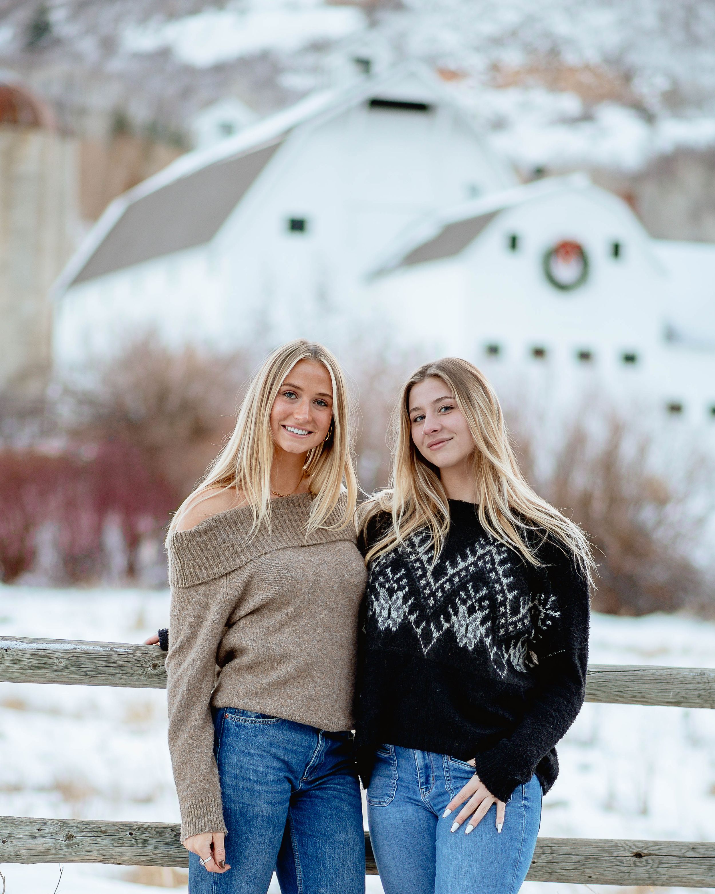 Two people in cozy sweaters standing by fence with white barn in snowy background.