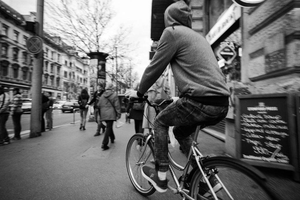 Urban street scene with cyclist riding past storefronts on a busy sidewalk.
