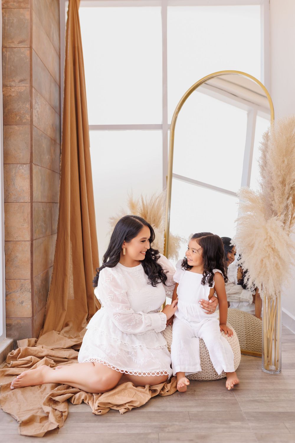 Mother and child in matching white dresses share tender moment on wooden floor.