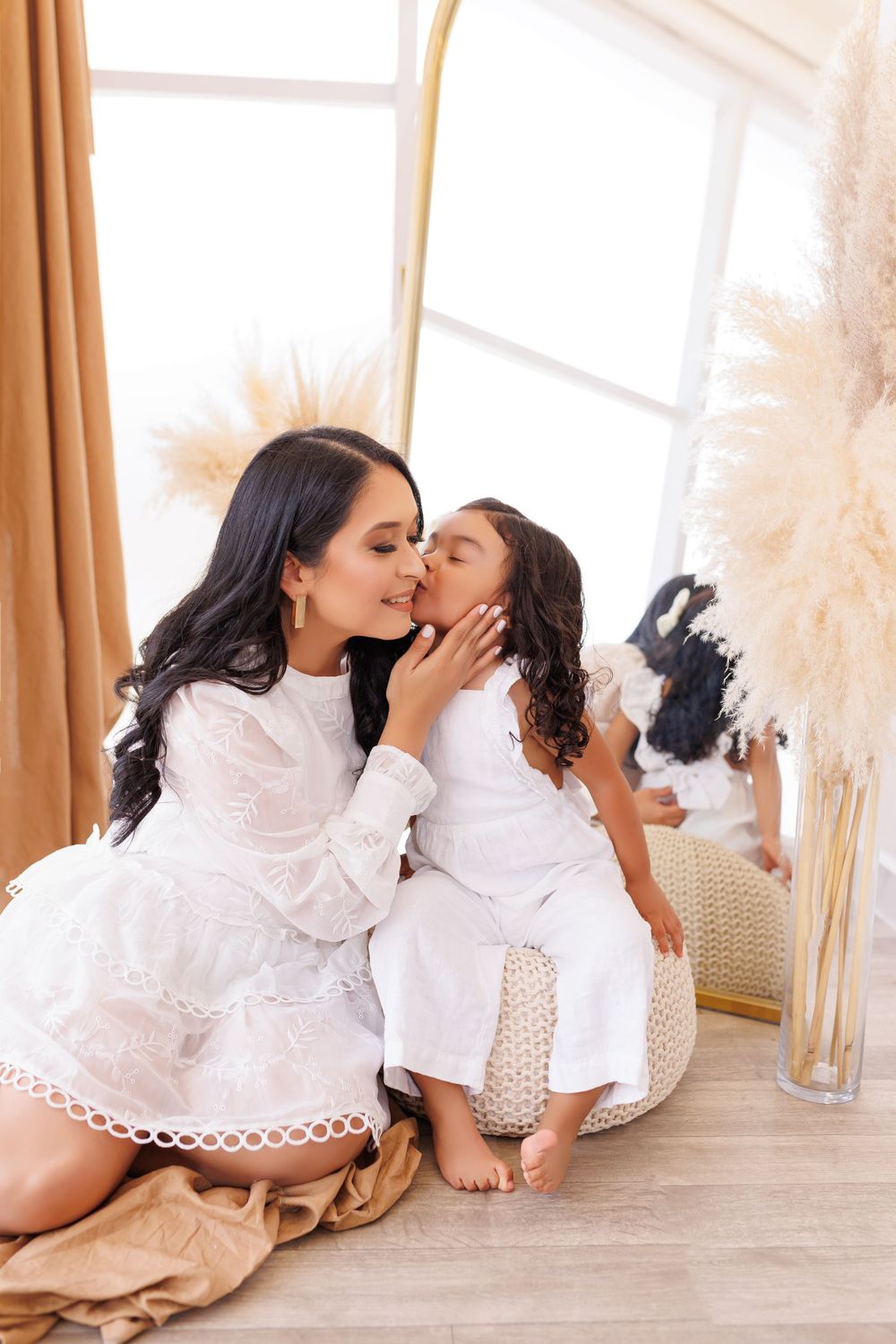 Mother and child wearing white lace dresses embrace on floor with pampas grass decor.