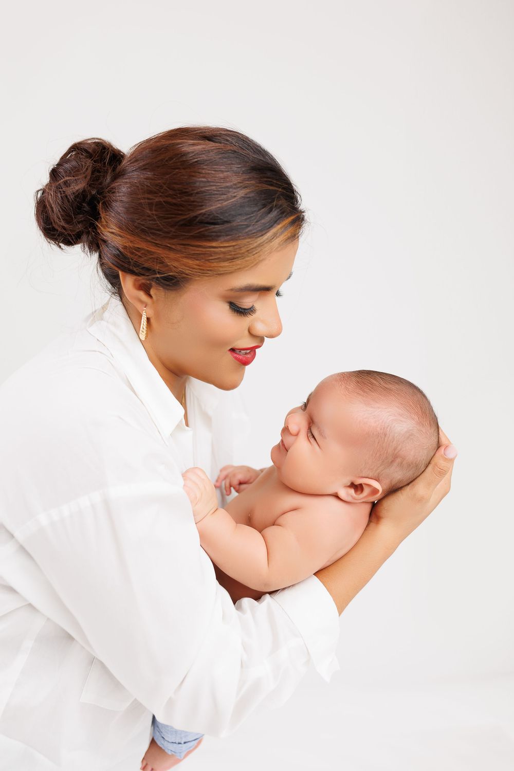 Woman in white shirt tenderly holding and looking down at newborn baby against white backdrop.