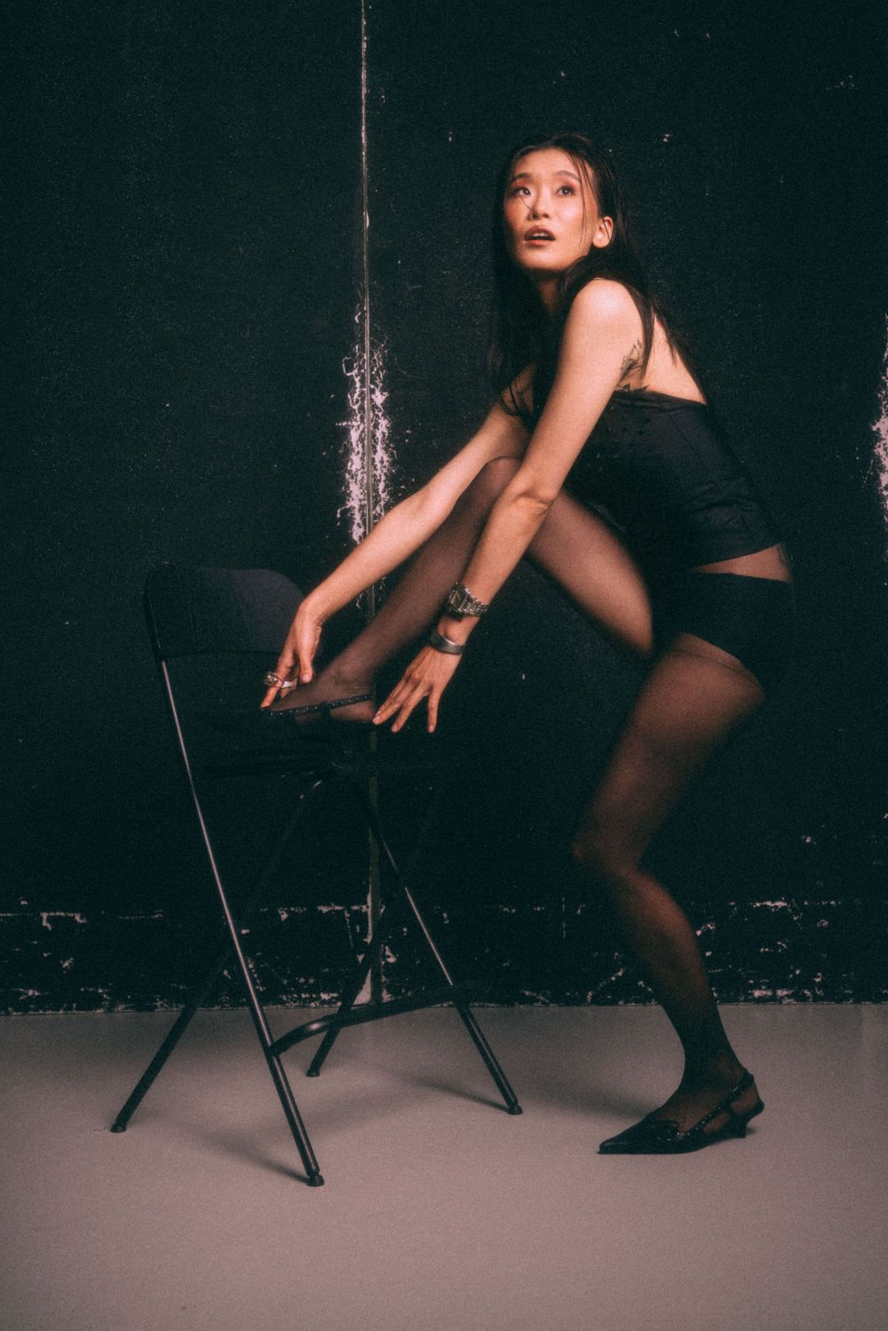 Dance pose against dark wall with black chair showing dynamic movement in black attire.