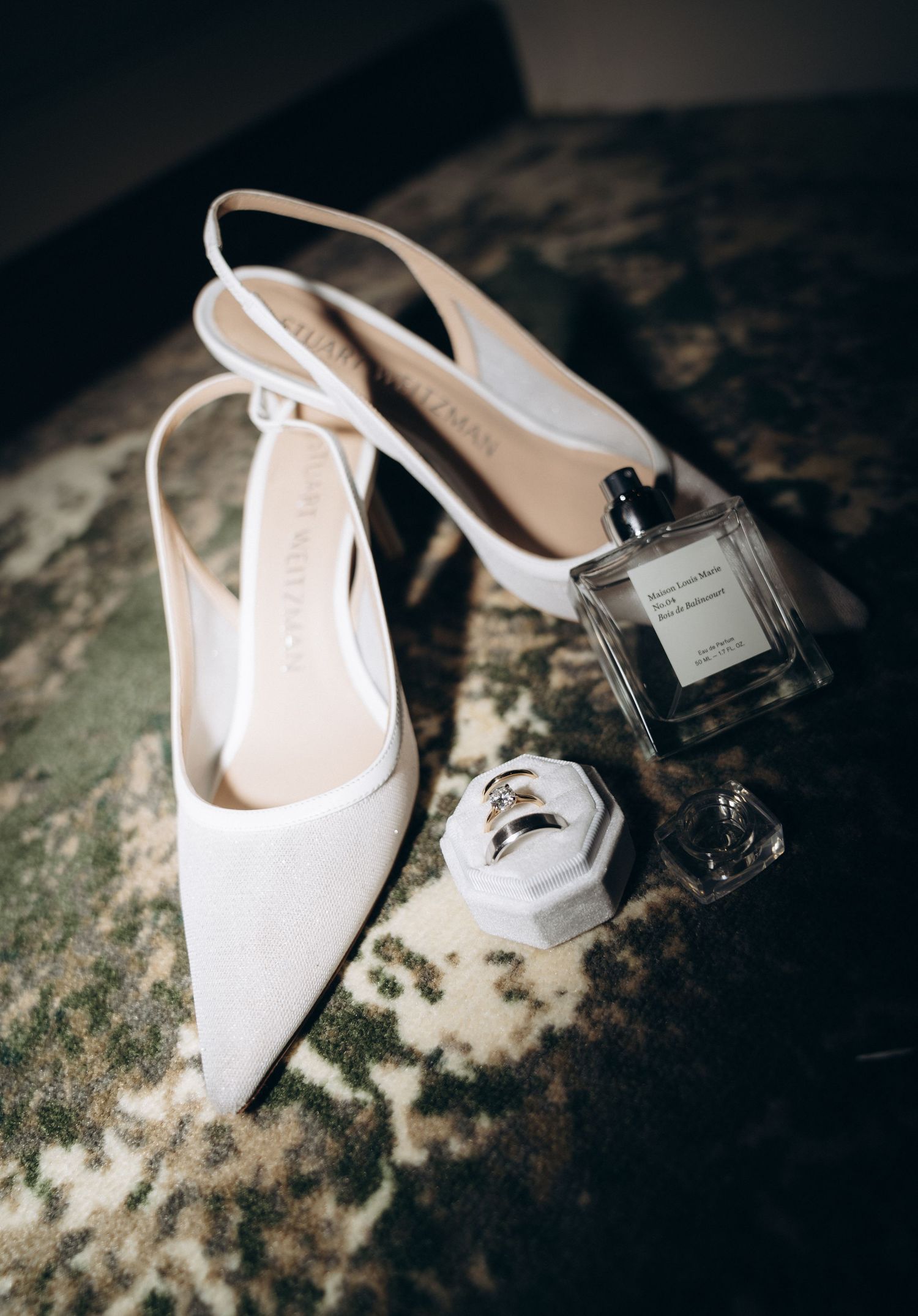 Elegant white slingback heels displayed with perfume bottles and accessories in moody lighting on textured surface.