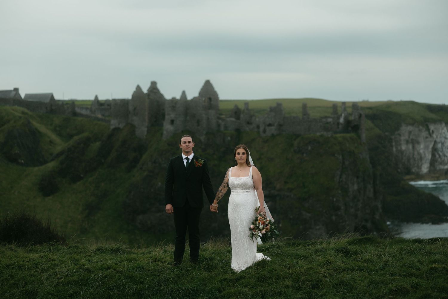 A couple poses for their wedding photos against the dramatic backdrop of Dunluce Castle ruins in Northern Ireland.