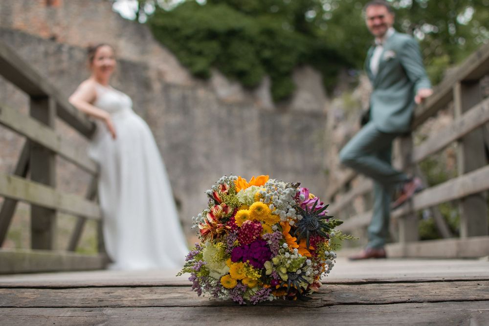 A colorful wildflower wedding bouquet in focus on wooden bridge with couple standing in soft focus background.