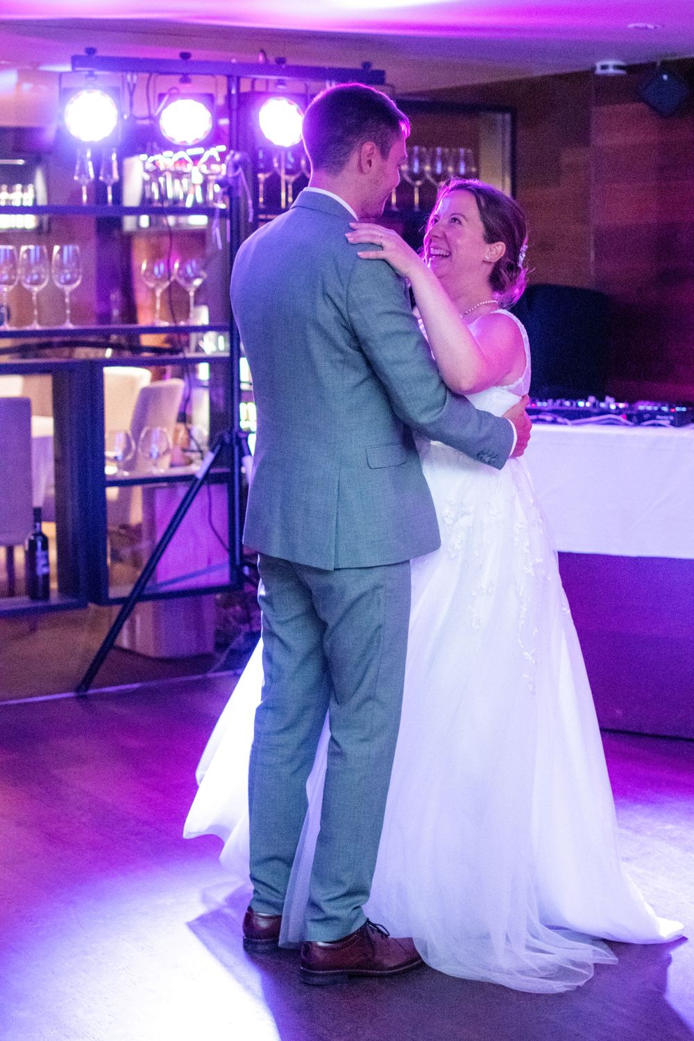 A couple shares their first dance at a wedding reception with purple mood lighting and a bar in the background.