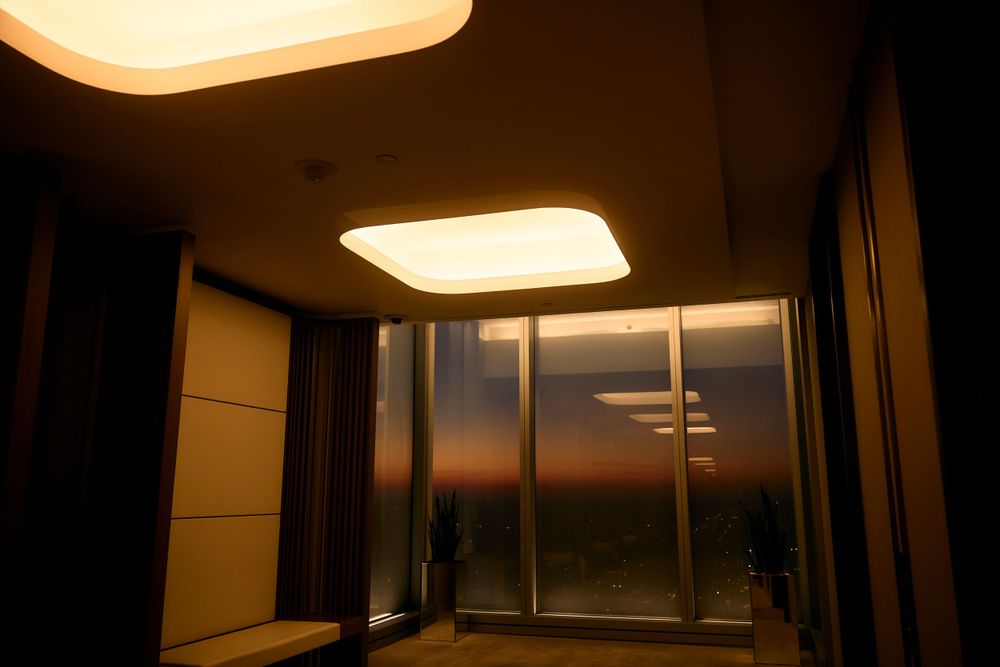 Modern hotel room at sunset with large illuminated ceiling panels and floor-to-ceiling windows.