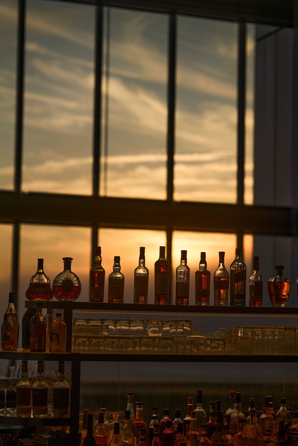 Backlit bar shelves displaying bottles of spirits against a dramatic sunset through window panels.