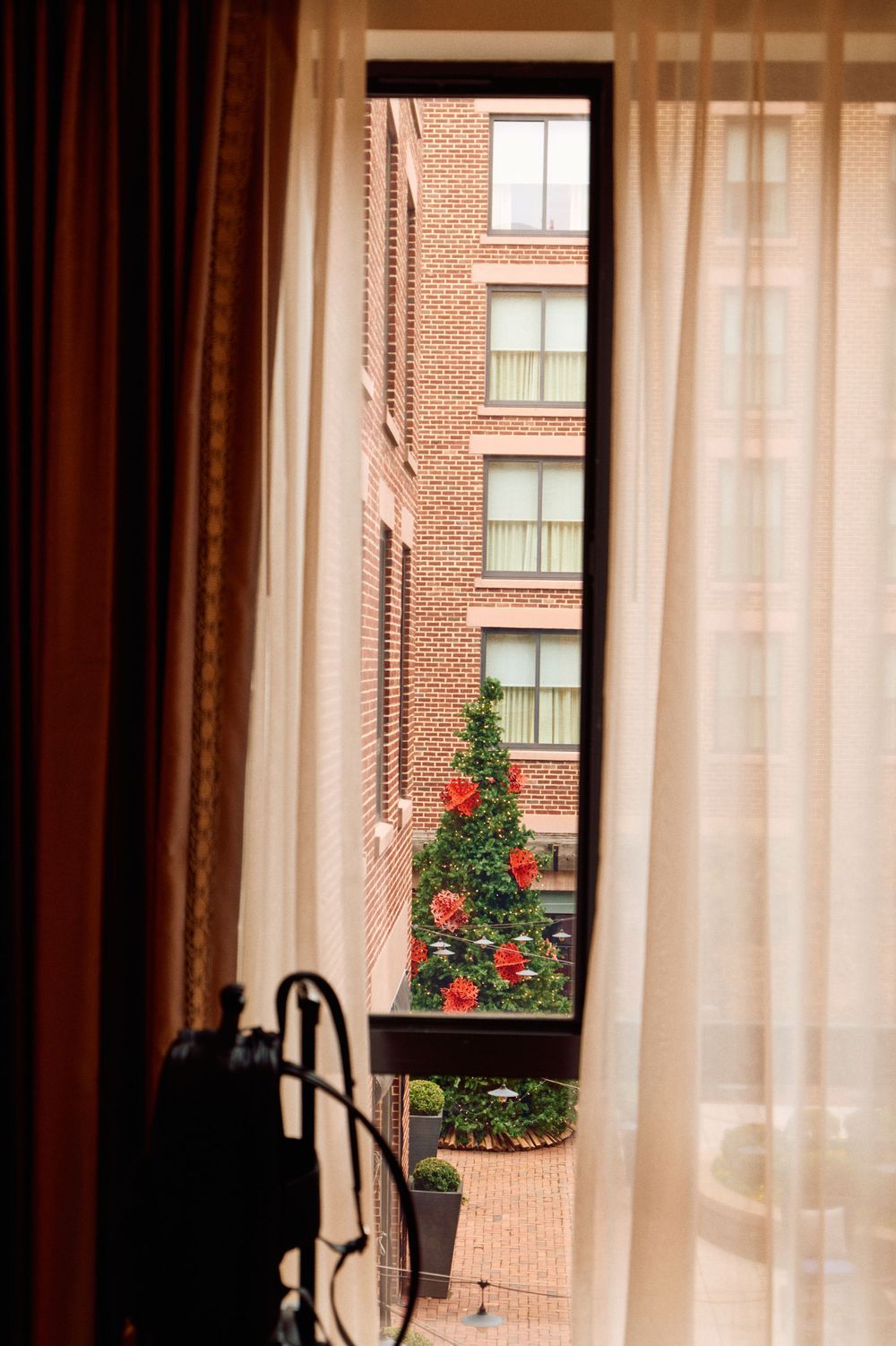 View through curtains of a red brick apartment building with flower planters visible on a sunny day.