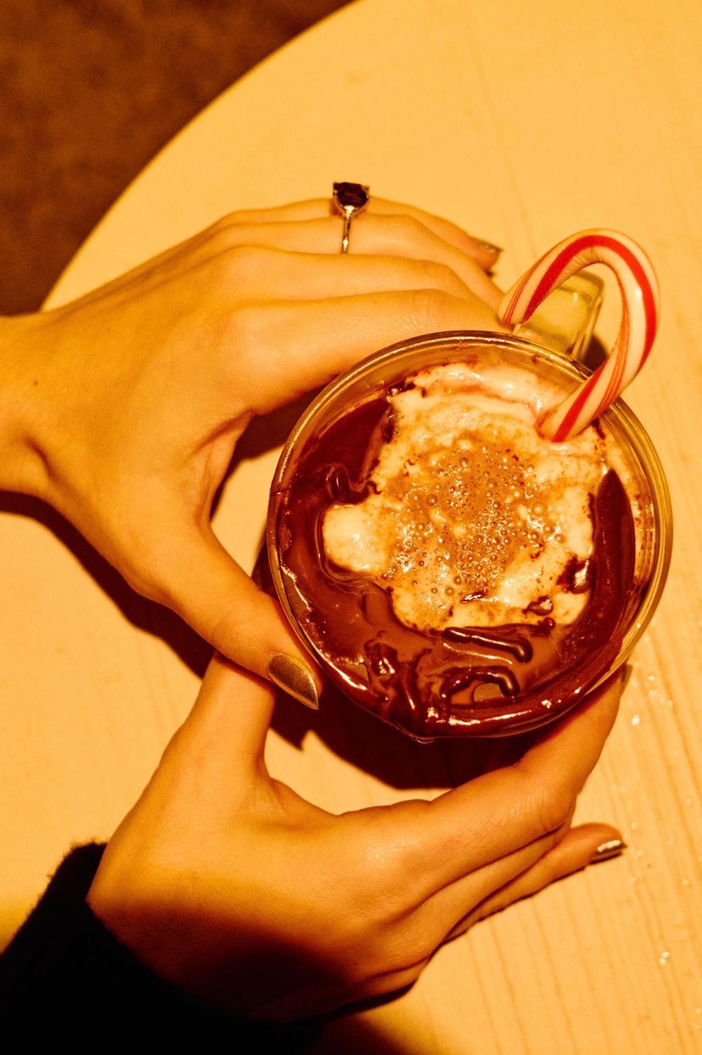 Hands holding festive chocolate drink topped with whipped cream and candy cane in warm lighting.