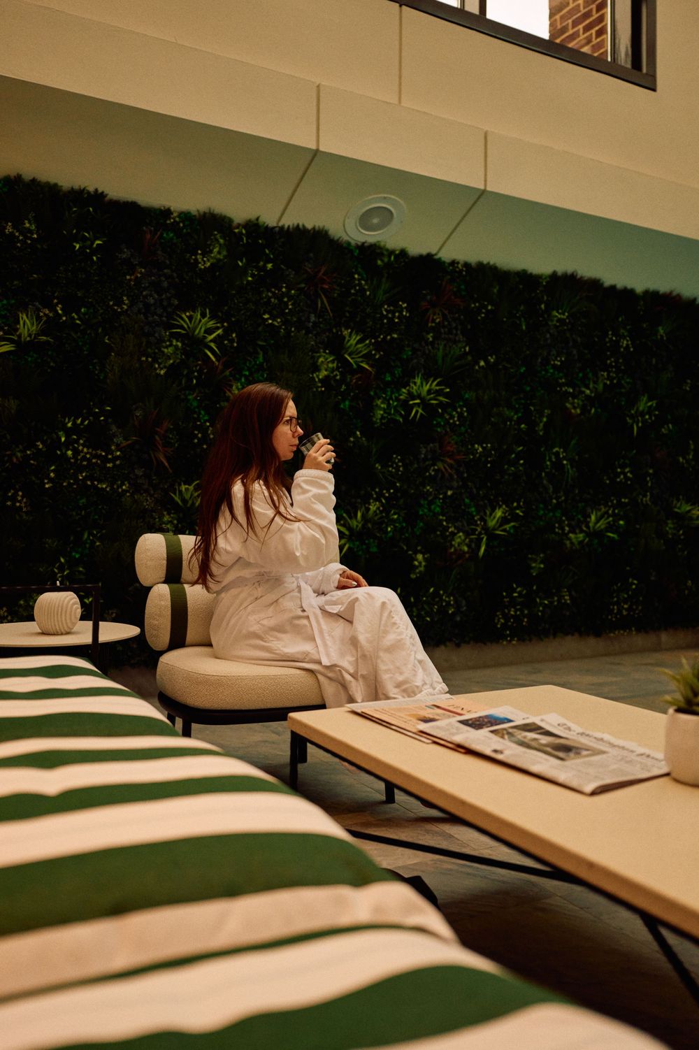 Person relaxing on lounge chair in white robe next to green wall with newspaper.