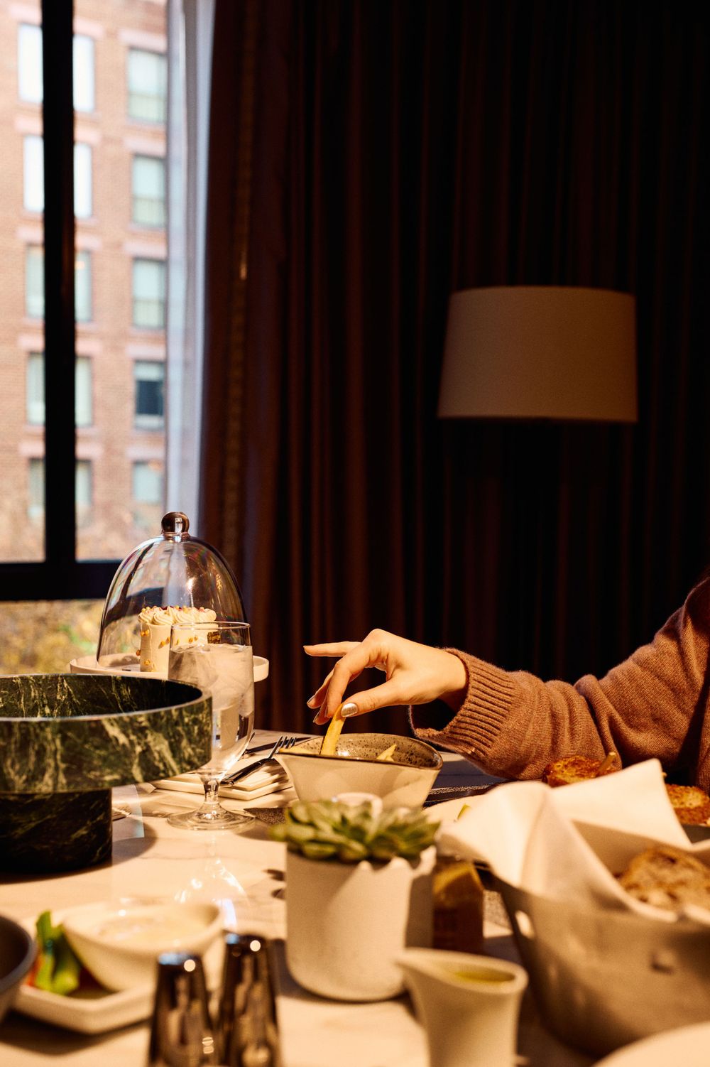 Hand reaching for food at elegant dining table with dark curtains and city view.