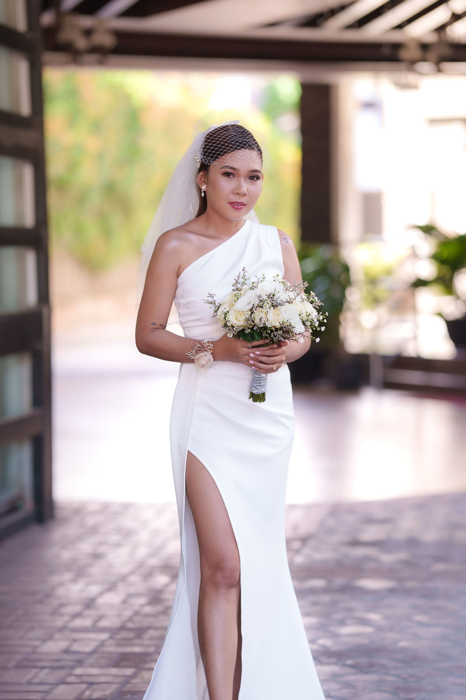 Stunning one-shoulder white wedding gown with thigh slit and delicate bouquet in modern venue.