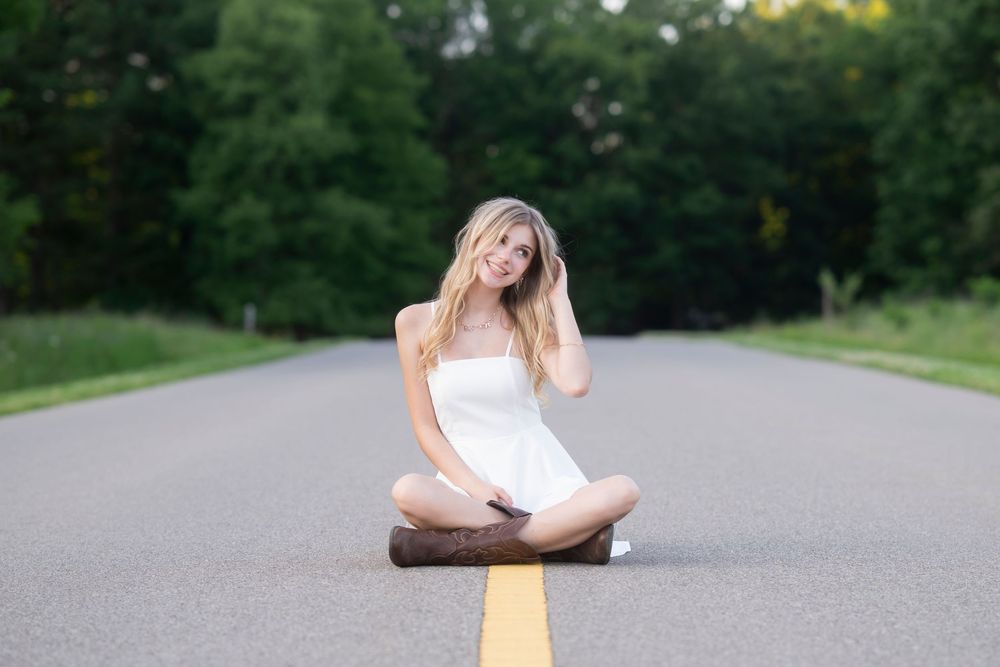 Person in white dress sits cross-legged on yellow road line during summer photoshoot.