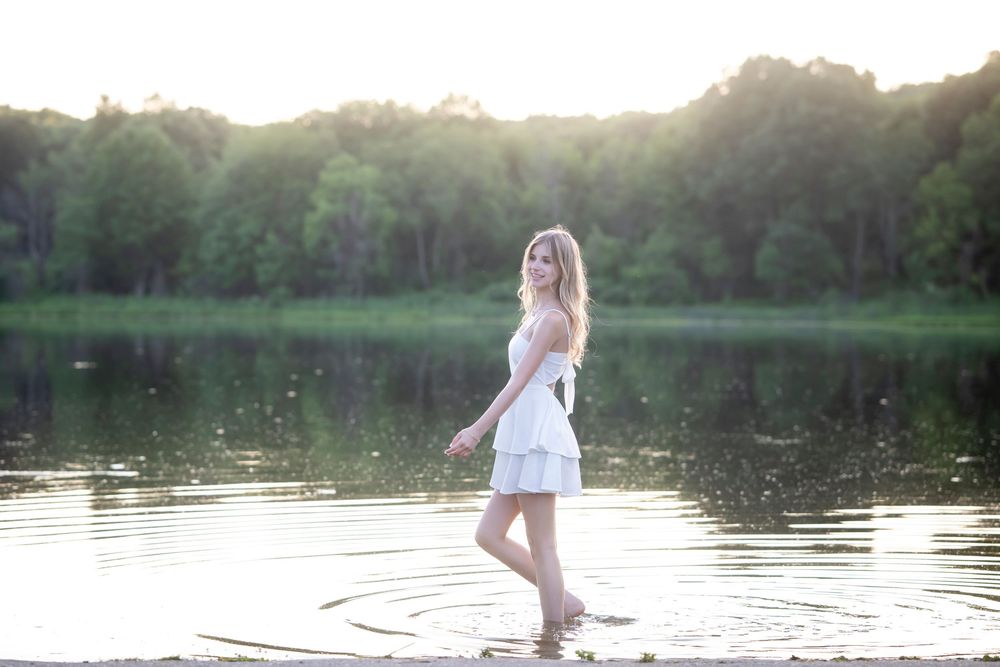 Person in a white dress walking along a serene lake at sunset creating ripples in the water.