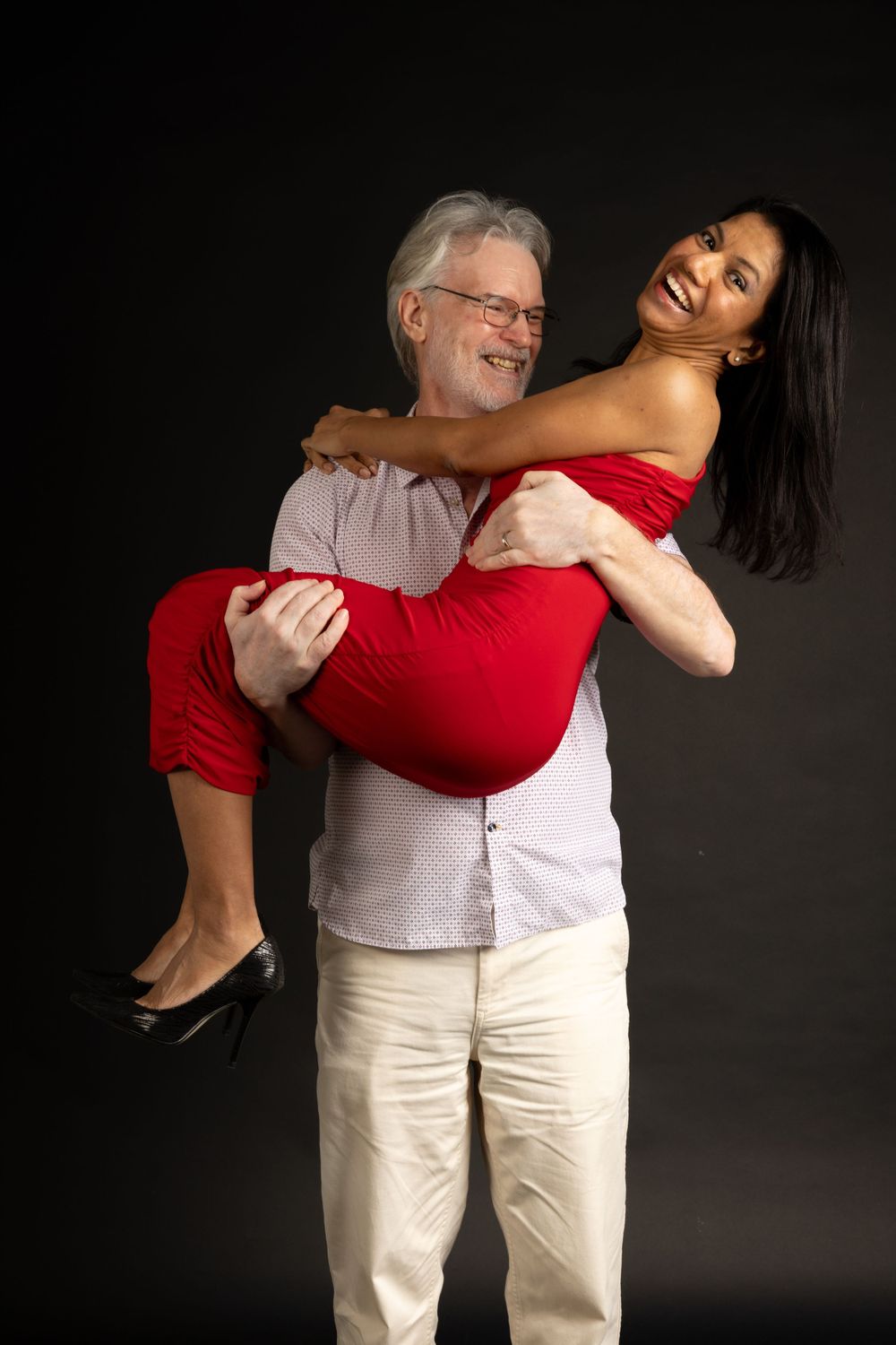 A couple poses playfully against a dark background, with one person in a red dress being held up by another in casual attire.