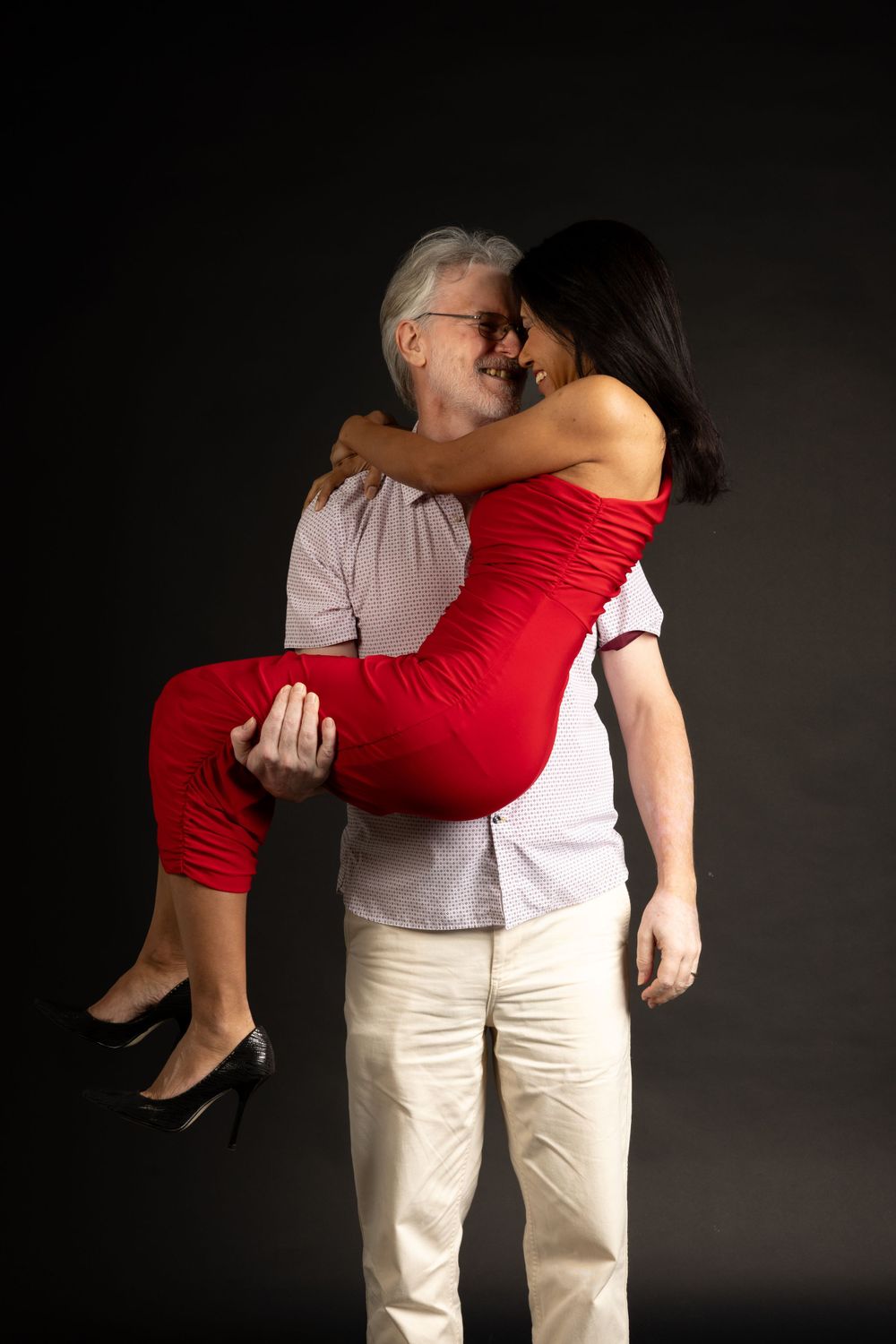 Couple in playful pose, man carrying woman in red dress against dark background.