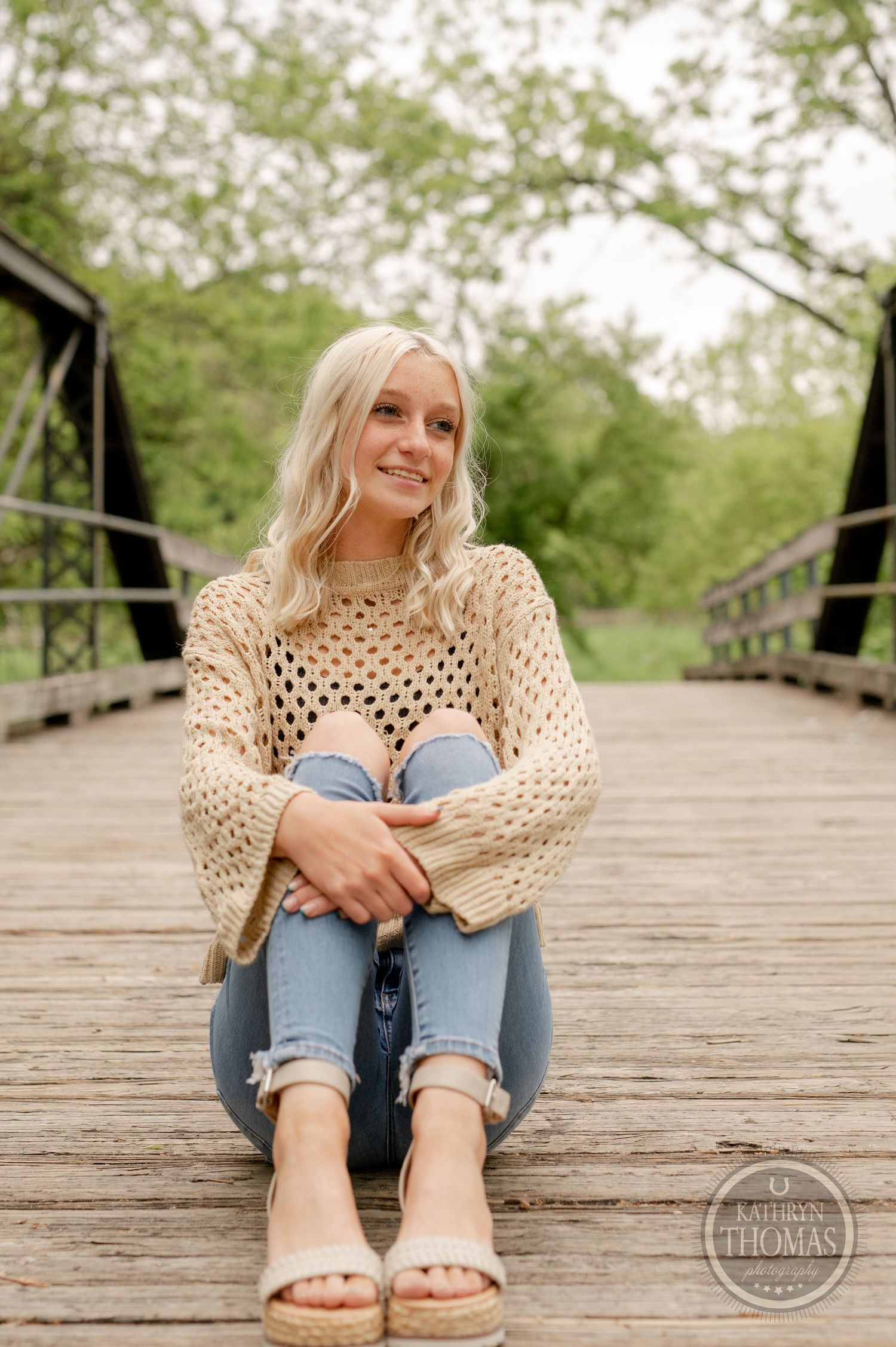Casual seated pose on wooden bridge wearing cream sweater and ripped jeans.
